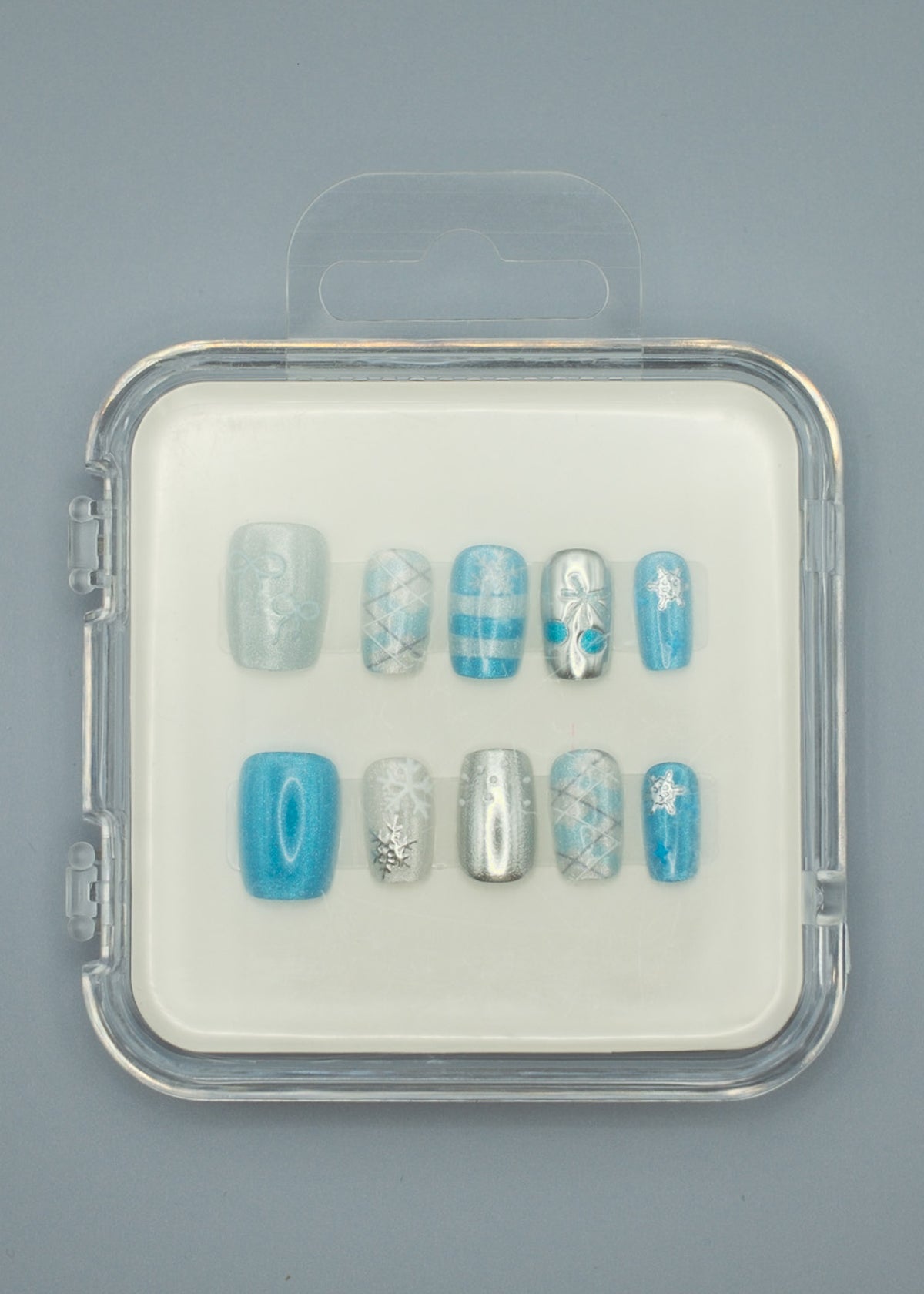Winter Wonderland by Mabel and Honey features reusable press-on nails in a clear case with silver, blue, and white snowflake, stripe, plaid, and solid designs—perfect for salon-quality manicures at home. Arranged neatly on a gray background.