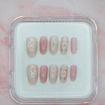 Bows 'n' Blushes by Mabel and Honey features reusable press-on nails in soft pink and beige with white bow and heart designs, neatly arranged in a clear plastic case against a pale pink marble background.