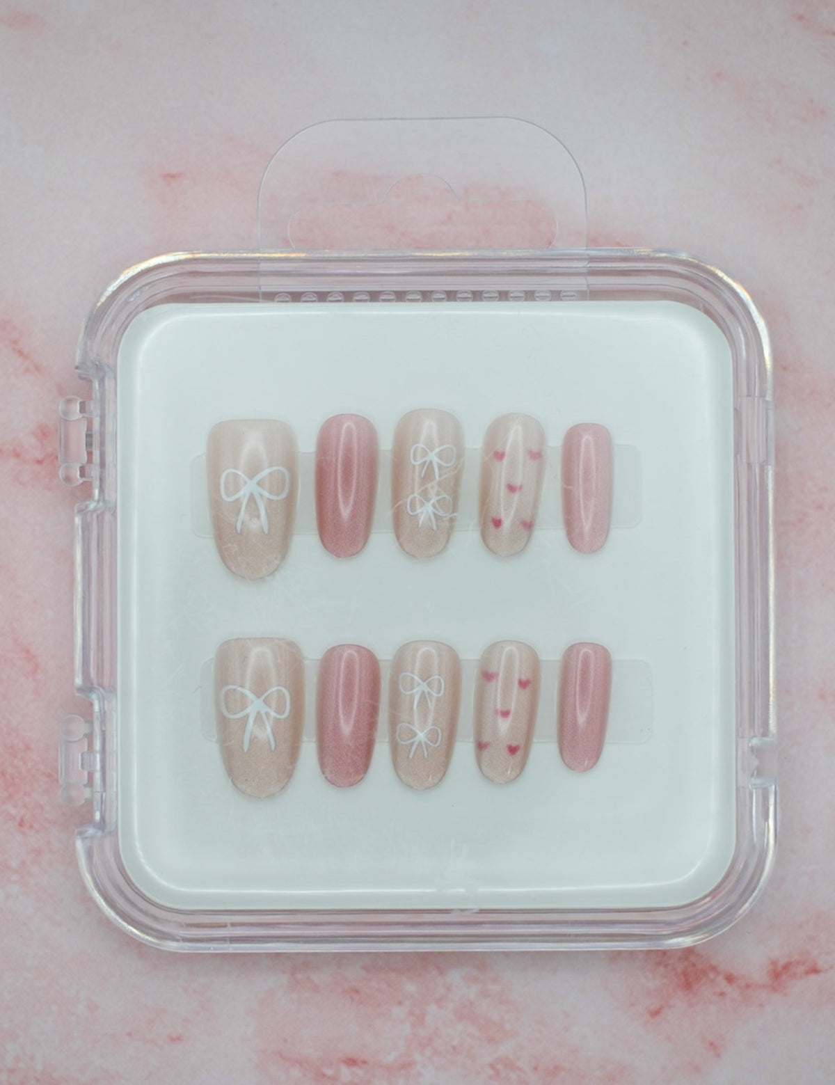 Bows 'n' Blushes by Mabel and Honey features reusable press-on nails in soft pink and beige with white bow and heart designs, neatly arranged in a clear plastic case against a pale pink marble background.