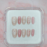 Bows 'n' Blushes by Mabel and Honey features reusable press-on nails in soft pink and beige with white bow and heart designs, neatly arranged in a clear plastic case against a pale pink marble background.