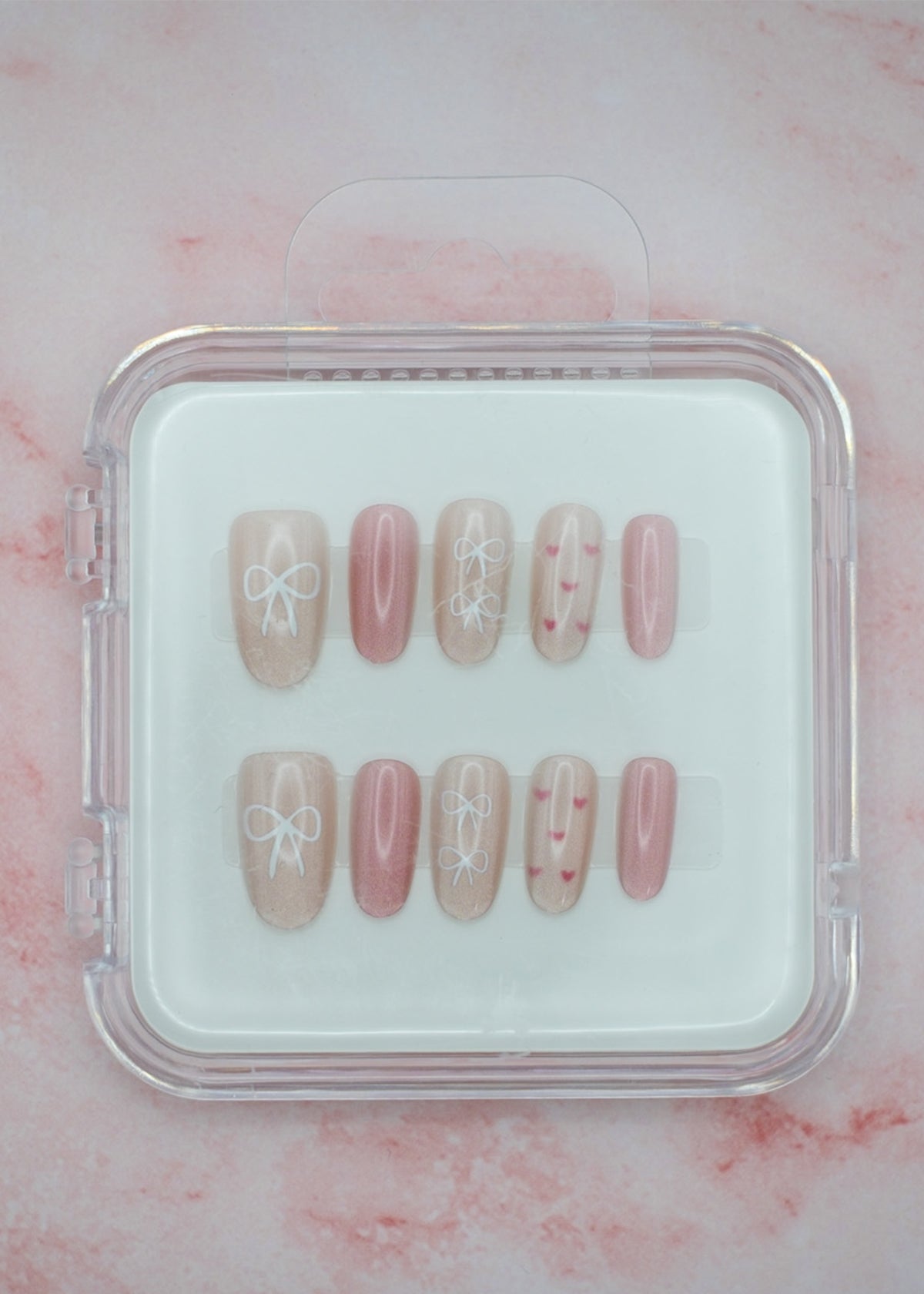 Bows 'n' Blushes by Mabel and Honey features reusable press-on nails in soft pink and beige with white bow and heart designs, neatly arranged in a clear plastic case against a pale pink marble background.
