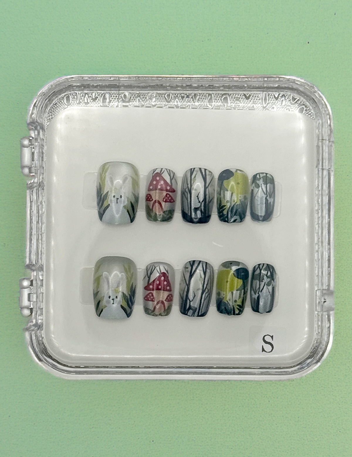 The Enchanted Bunny Acrylic Nails by Mabel and Honey are reusable and come in a clear case, featuring hand-painted white flowers, red mushrooms, and green-gray foliage on a mint green base for a salon-quality manicure.