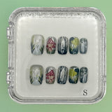 The Enchanted Bunny Acrylic Nails by Mabel and Honey are reusable and come in a clear case, featuring hand-painted white flowers, red mushrooms, and green-gray foliage on a mint green base for a salon-quality manicure.