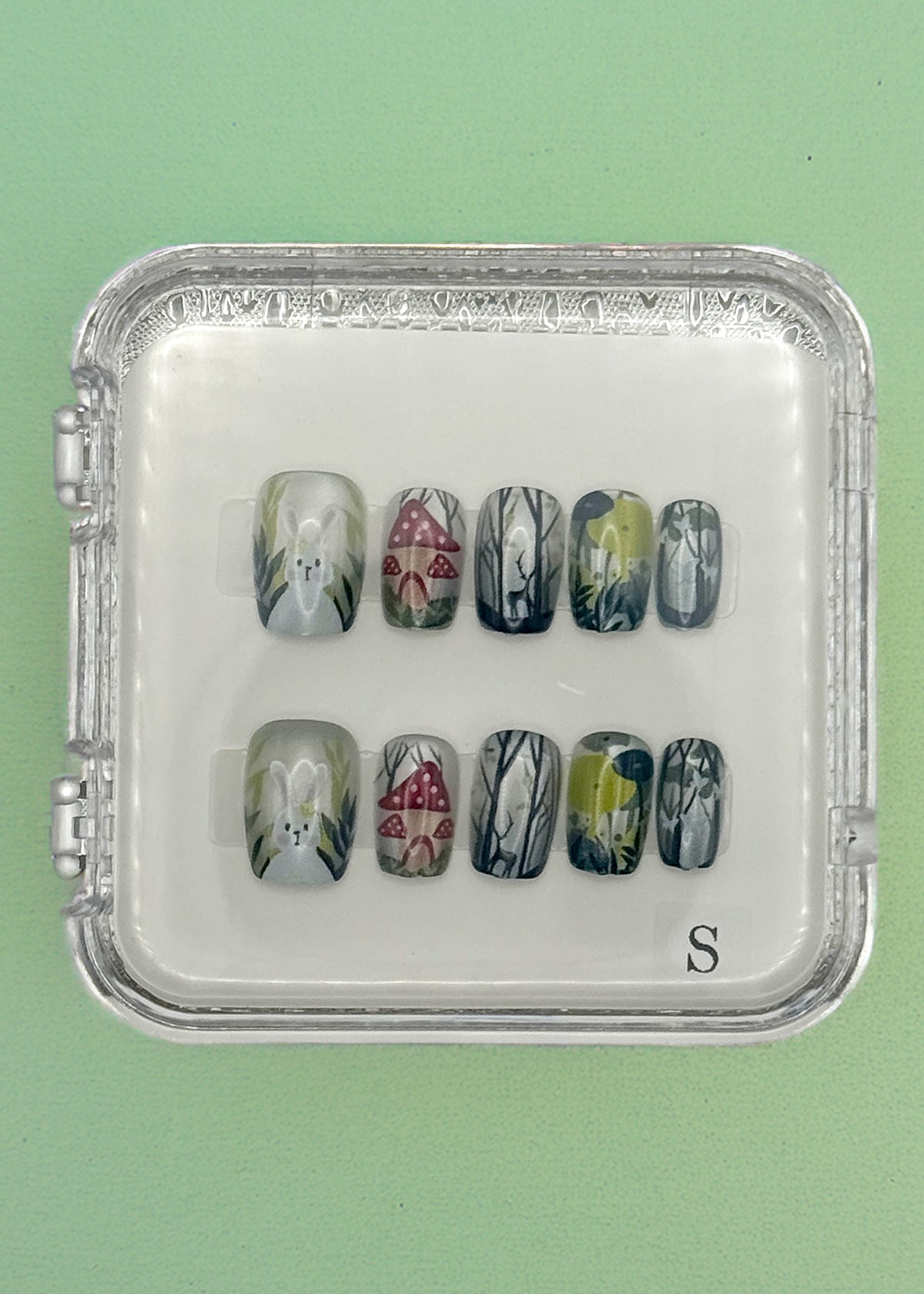 The Enchanted Bunny Acrylic Nails by Mabel and Honey are reusable and come in a clear case, featuring hand-painted white flowers, red mushrooms, and green-gray foliage on a mint green base for a salon-quality manicure.
