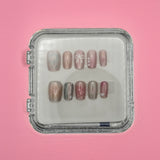 Wish You Well Acrylic Nails by Mabel and Honey come in a clear square case, featuring a shiny nude design with white lines, bows, and small flowers for a salon-quality manicure at home. Displayed on a light pink background.