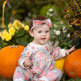 A baby in the Mabel and Honey Eliza Long Sleeve Floral Ribbed Trim Gathered Waist Bubble Romper sits barefoot on the grass between pumpkins, surrounded by blooming flowers and greenery, smiling at the camera.