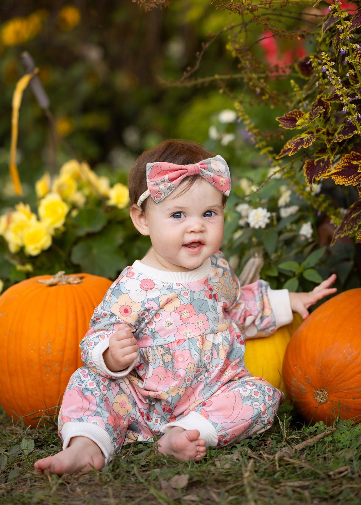 A baby in the Mabel and Honey Eliza Long Sleeve Floral Ribbed Trim Gathered Waist Bubble Romper sits barefoot on the grass between pumpkins, surrounded by blooming flowers and greenery, smiling at the camera.