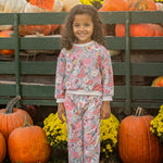 A young girl with curly hair, dressed in the Mabel and Honey Eliza Long Sleeve Floral Sweatshirt and Jogger Set made from soft knit fabric, pairs her outfit with white sneakers as she smiles in front of pumpkins, vibrant yellow flowers, and a wooden display at an outdoor pumpkin patch.