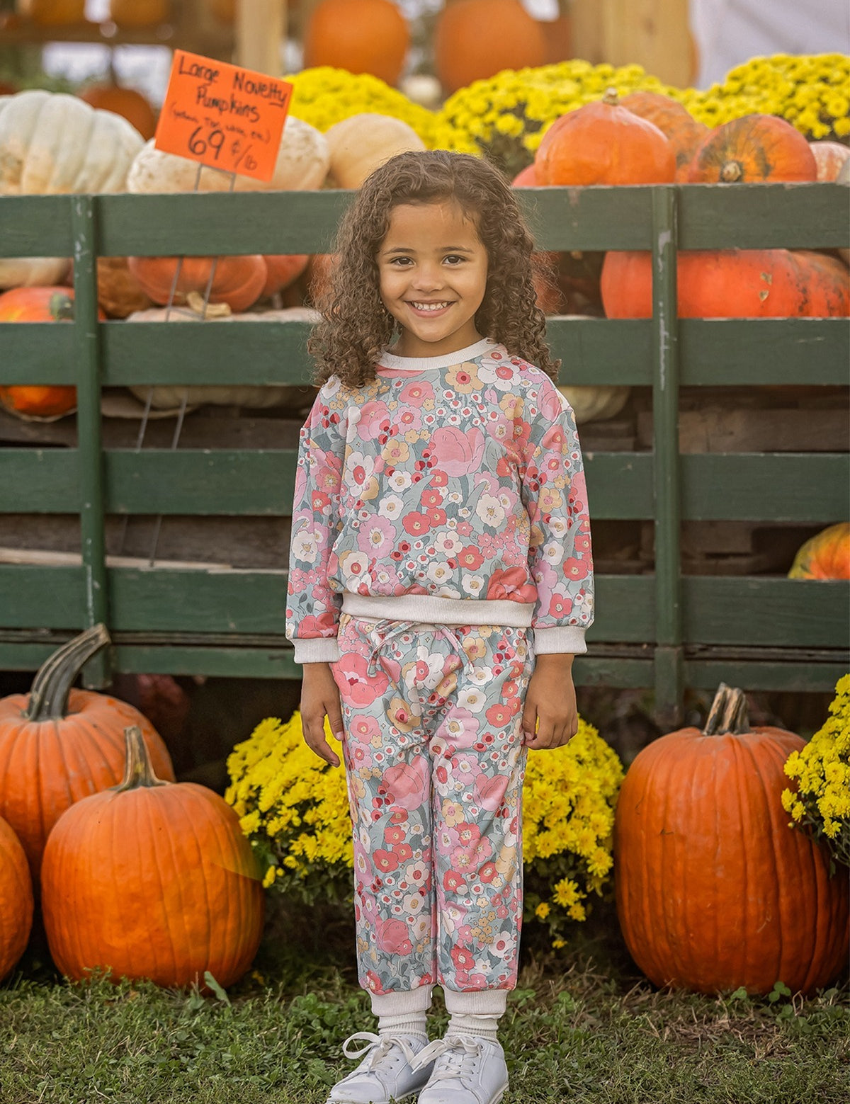 A young girl with curly hair, dressed in the Mabel and Honey Eliza Long Sleeve Floral Sweatshirt and Jogger Set made from soft knit fabric, pairs her outfit with white sneakers as she smiles in front of pumpkins, vibrant yellow flowers, and a wooden display at an outdoor pumpkin patch.