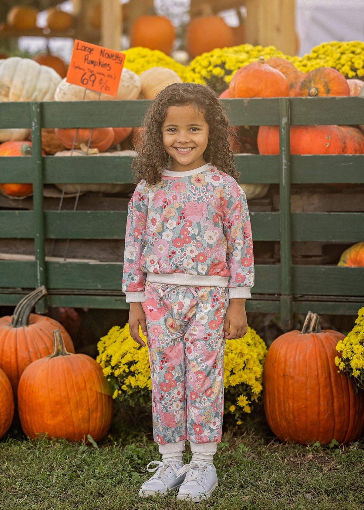 A young girl with curly hair, dressed in the Mabel and Honey Eliza Long Sleeve Floral Sweatshirt and Jogger Set made from soft knit fabric, pairs her outfit with white sneakers as she smiles in front of pumpkins, vibrant yellow flowers, and a wooden display at an outdoor pumpkin patch.