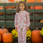 A young girl with curly hair smiles among orange pumpkins and yellow flowers, wearing the Eliza Long Sleeve Floral Sweatshirt and Jogger Set by Mabel and Honey. Green wooden crates filled with pumpkins are in the background.