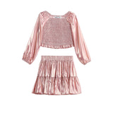 The Rosy Glow Two Piece Set by Mabel + Honey features a shiny pink metallic rose gold long-sleeve smocked crop top with gathered detailing, paired with a matching tiered skirt adorned with playful ruffles.