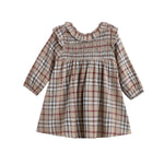 The Mabel + Honey Chestnut Dress is a long-sleeved, A-line baby dress featuring a plaid pattern in beige, red, and gray tones, ruffled shoulders, and a hand-smocked bodice. Crafted from a soft cotton blend flannel, it is displayed on a white background.