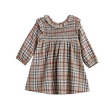 The Mabel + Honey Chestnut Dress is a long-sleeved, A-line baby dress featuring a plaid pattern in beige, red, and gray tones, ruffled shoulders, and a hand-smocked bodice. Crafted from a soft cotton blend flannel, it is displayed on a white background.