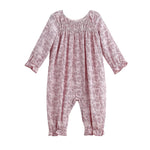 The Loretta Romper Long Sleeve Floral Smocked Straight Leg Romper by Mabel + Honey is shown on a white background, featuring a light pink fabric with a subtle floral print, smocked yoke, gathered neckline, and ruffled cuffs at the sleeves and ankles.