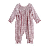 The Loretta Romper Long Sleeve Floral Smocked Straight Leg Romper by Mabel + Honey is shown on a white background, featuring a light pink fabric with a subtle floral print, smocked yoke, gathered neckline, and ruffled cuffs at the sleeves and ankles.