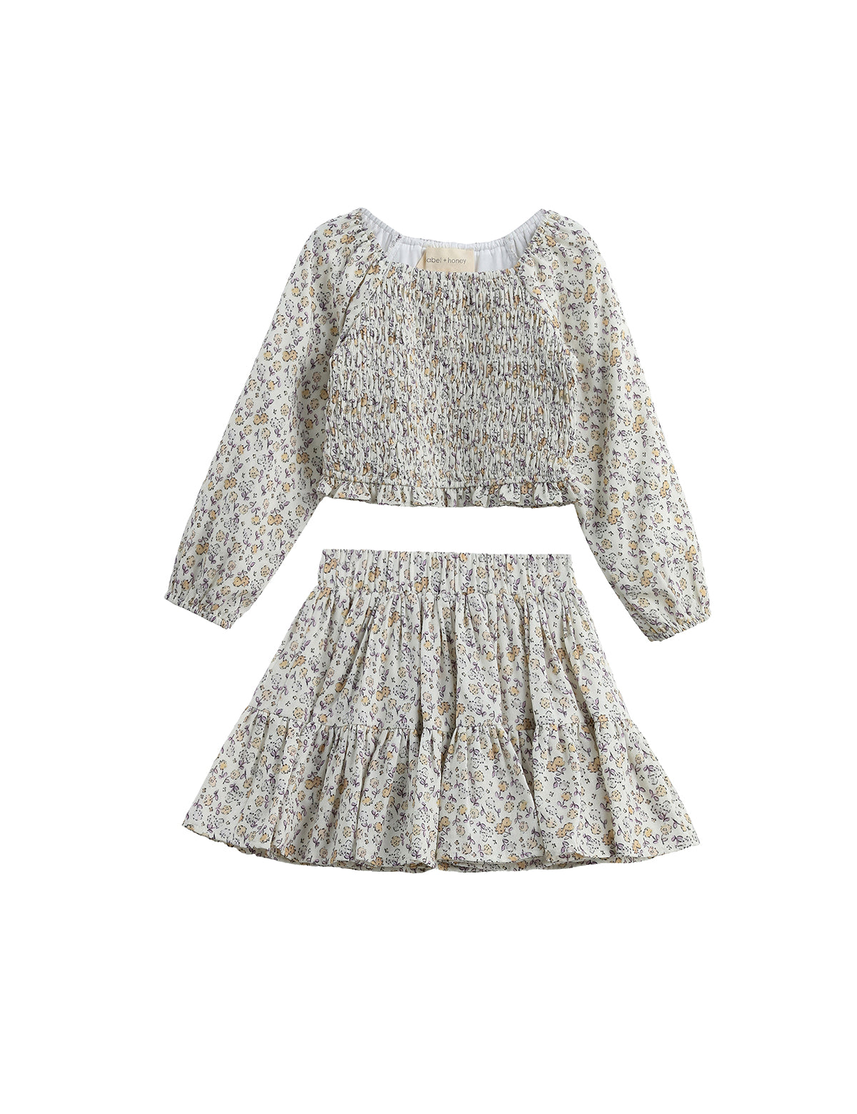The Wildflower Dream Two Piece Set by Mabel + Honey showcases a long sleeve floral smocked top paired with a matching tiered mini skirt, both crafted from lightweight fabric adorned with a delicate small flower print. This charming set is displayed against a white background.