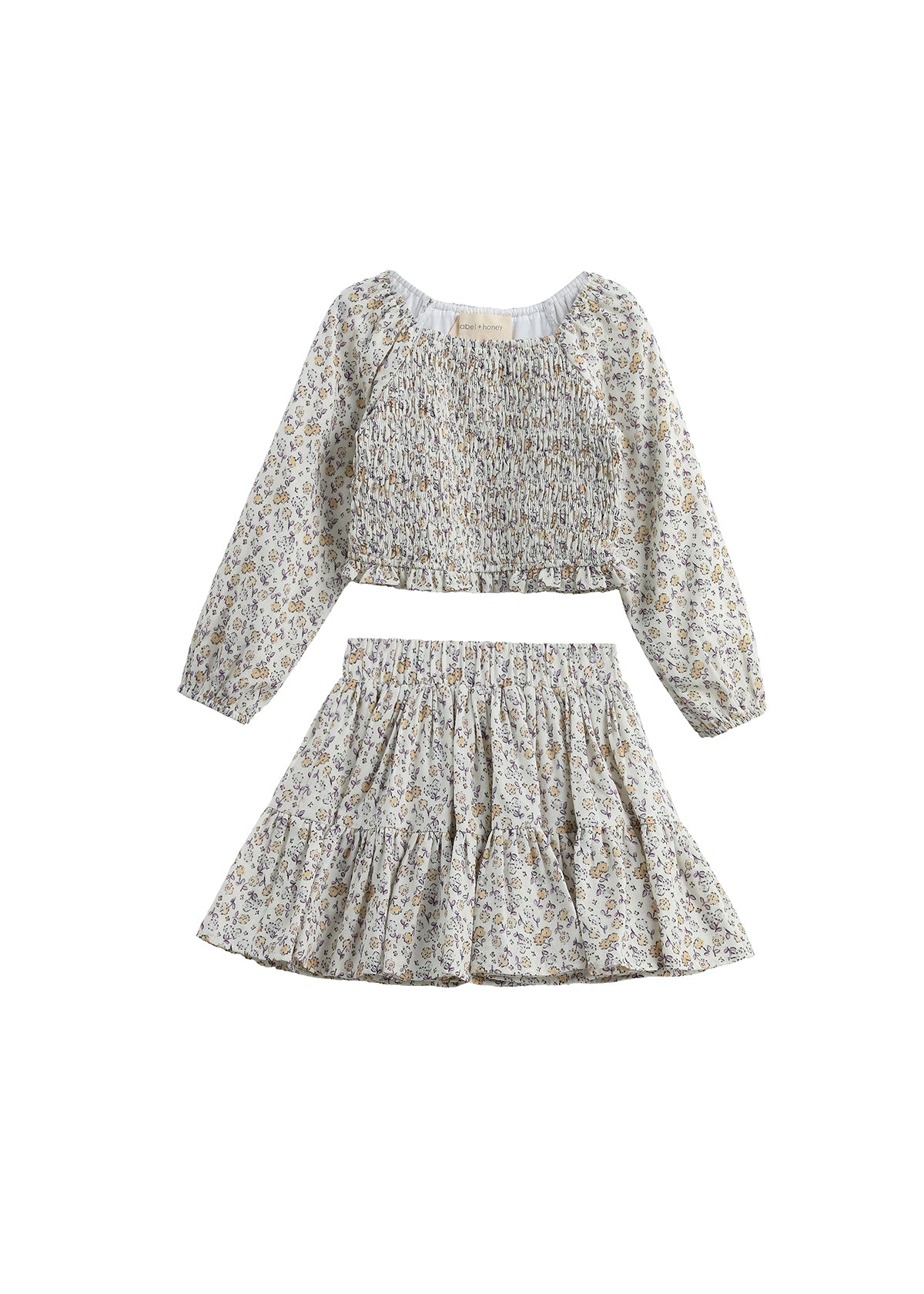 The Wildflower Dream Two Piece Set by Mabel + Honey showcases a long sleeve floral smocked top paired with a matching tiered mini skirt, both crafted from lightweight fabric adorned with a delicate small flower print. This charming set is displayed against a white background.