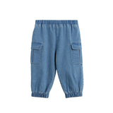 The Michael Pants Solid Chambray Cargo Jogger Style Elastic Cuff Pants by Mabel + Honey are girls’ light denim joggers with a classic cargo design, featuring an elastic waistband and cuffs for comfort. These chambray pants include two large side flap pockets for added utility and are shown laid flat on a white background.