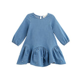 The Mabel + Honey Denim Doll Dress is a long-sleeve, solid chambray dress for children, featuring a dropwaist silhouette with a flared, ruffled hem accented by delicate white pearl embellishments and a classic round neckline.