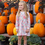 A young girl wearing the Eliza Long Sleeve Floral Ribbed Trim Empire Waist Dress by Mabel and Honey stands smiling in front of stacked pumpkins and leafy greens outside a store, with orange price signs visible in the background.