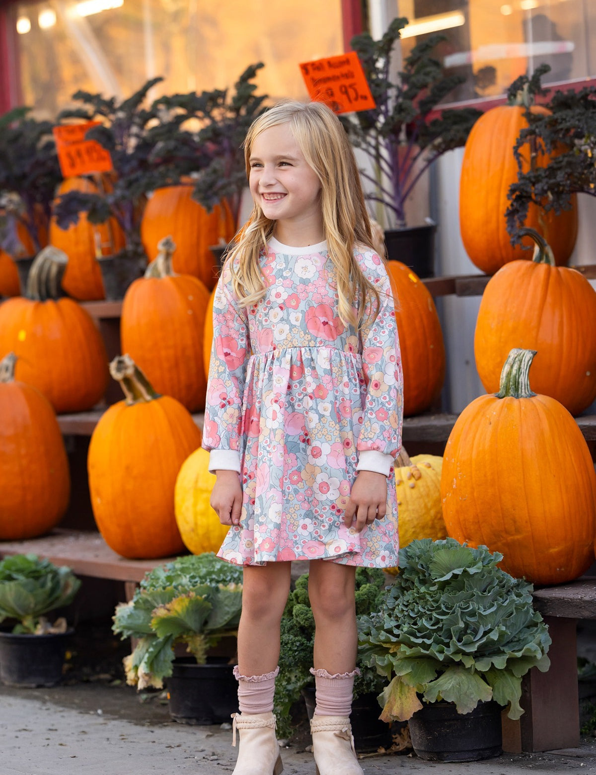 A young girl wearing the Eliza Long Sleeve Floral Ribbed Trim Empire Waist Dress by Mabel and Honey stands smiling in front of stacked pumpkins and leafy greens outside a store, with orange price signs visible in the background.