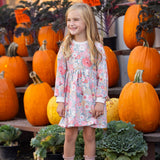 A young girl wearing the Eliza Long Sleeve Floral Ribbed Trim Empire Waist Dress by Mabel and Honey stands smiling in front of stacked pumpkins and leafy greens outside a store, with orange price signs visible in the background.