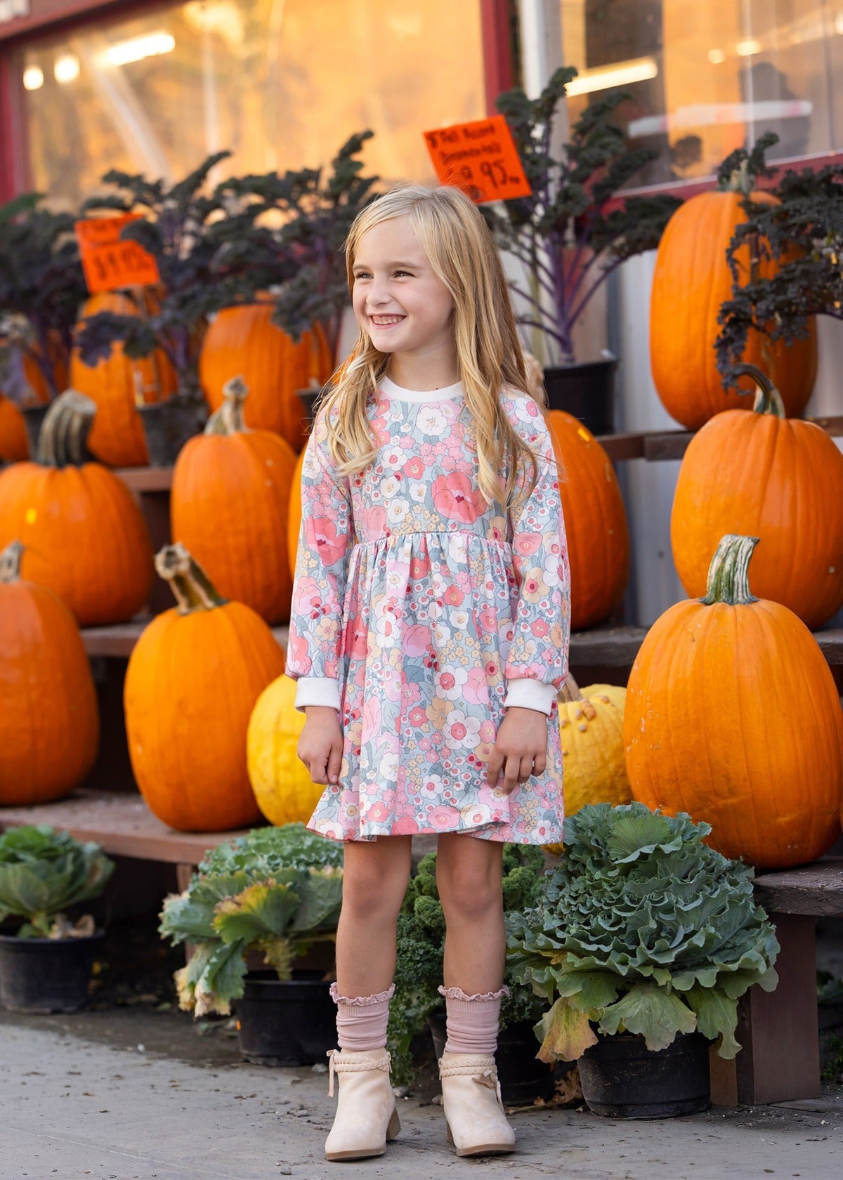 A young girl wearing the Eliza Long Sleeve Floral Ribbed Trim Empire Waist Dress by Mabel and Honey stands smiling in front of stacked pumpkins and leafy greens outside a store, with orange price signs visible in the background.