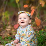 A smiling baby wearing the Mabel and Honey Ivy Bloom Long Sleeve Floral Ribbed Trim Straight Leg Romper with a matching headband sits on the grass outdoors, surrounded by autumn leaves and greenery.