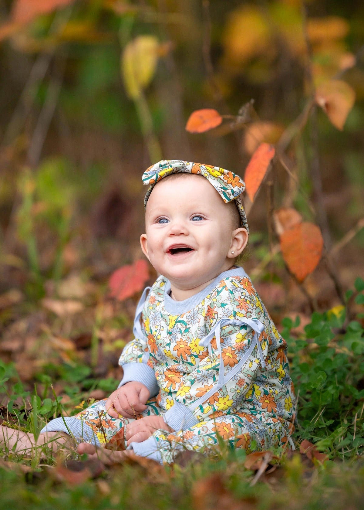 A smiling baby wearing the Mabel and Honey Ivy Bloom Long Sleeve Floral Ribbed Trim Straight Leg Romper with a matching headband sits on the grass outdoors, surrounded by autumn leaves and greenery.