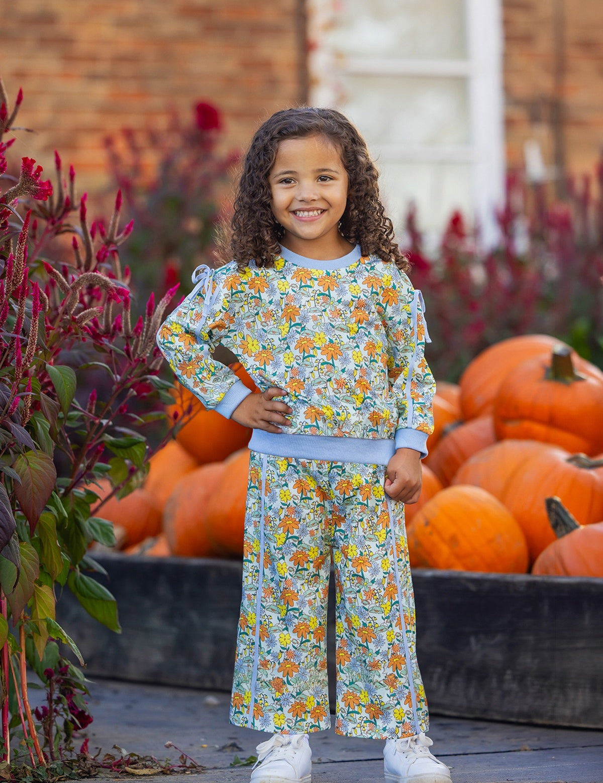 A young child with curly hair smiles, standing in front of a pile of pumpkins. They wear the Ivy Bloom Long Sleeve Floral Sweatshirt and Wide Leg Pant Set from Mabel and Honey, featuring blue trim and wide-leg pants, paired with white sneakers. The background includes red flowers and a brick building.