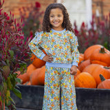 A young child with curly hair smiles, standing in front of a pile of pumpkins. They wear the Ivy Bloom Long Sleeve Floral Sweatshirt and Wide Leg Pant Set from Mabel and Honey, featuring blue trim and wide-leg pants, paired with white sneakers. The background includes red flowers and a brick building.