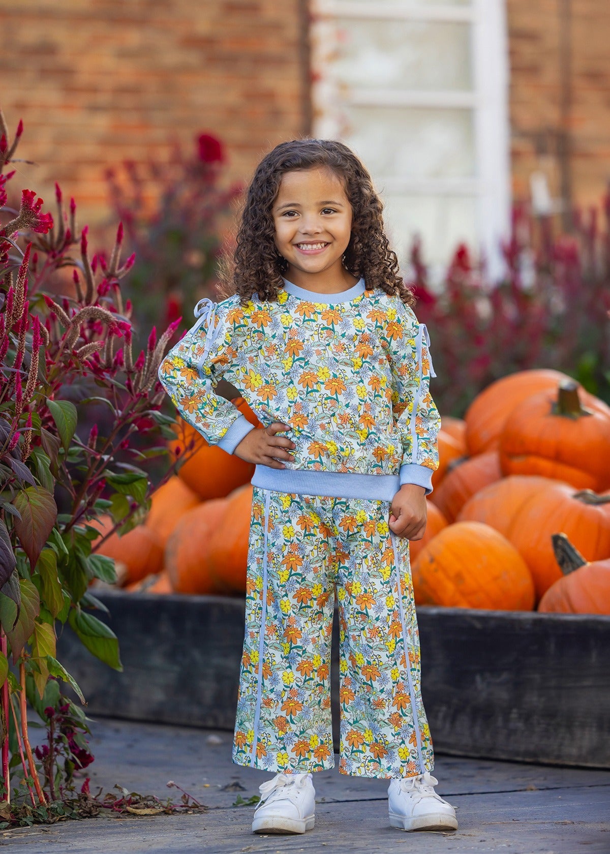 A young child with curly hair smiles, standing in front of a pile of pumpkins. They wear the Ivy Bloom Long Sleeve Floral Sweatshirt and Wide Leg Pant Set from Mabel and Honey, featuring blue trim and wide-leg pants, paired with white sneakers. The background includes red flowers and a brick building.