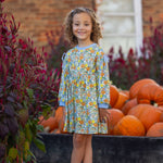 A young girl wearing the Mabel and Honey Ivy Bloom Long Sleeve Floral Ribbed Trim Bow Tie Dress stands smiling near a flower bed and a wooden crate filled with pumpkins, with a brick building and window in the background.