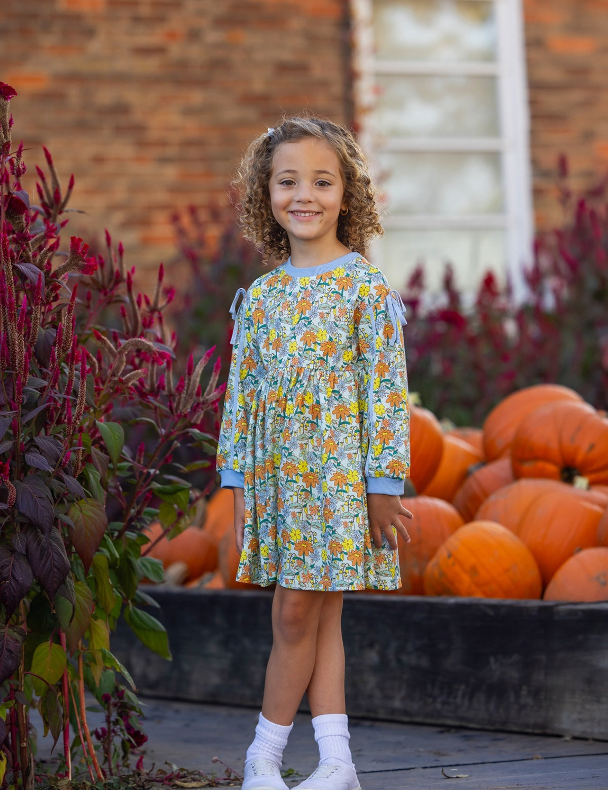 A young girl wearing the Mabel and Honey Ivy Bloom Long Sleeve Floral Ribbed Trim Bow Tie Dress stands smiling near a flower bed and a wooden crate filled with pumpkins, with a brick building and window in the background.