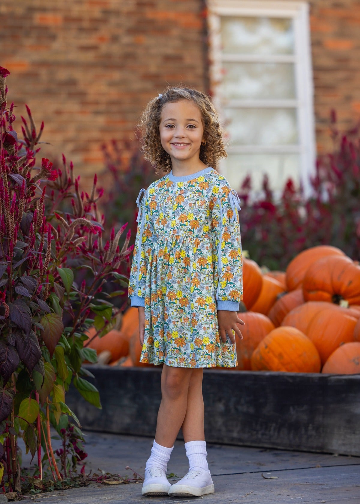 A young girl wearing the Mabel and Honey Ivy Bloom Long Sleeve Floral Ribbed Trim Bow Tie Dress stands smiling near a flower bed and a wooden crate filled with pumpkins, with a brick building and window in the background.