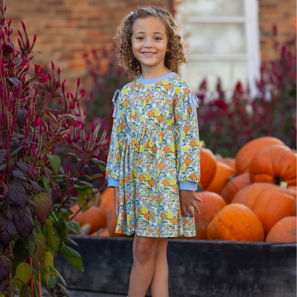 A young girl with curly hair, wearing the Mabel and Honey Ivy Bloom Long Sleeve Floral Ribbed Trim Bow Tie Dress, stands smiling in front of a pile of pumpkins and tall red plants, with a brick building and window in the background.