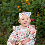 A smiling baby in the Mabel and Honey Floral Flutter Long Sleeve Ruffle Shoulder Bubble Romper, featuring a soft stretch knit fabric with charming floral patterns and a matching bow, sits on green grass as vibrant orange and yellow flowers bloom in the background. The adorable bubble leg design adds extra charm to this delightful scene.