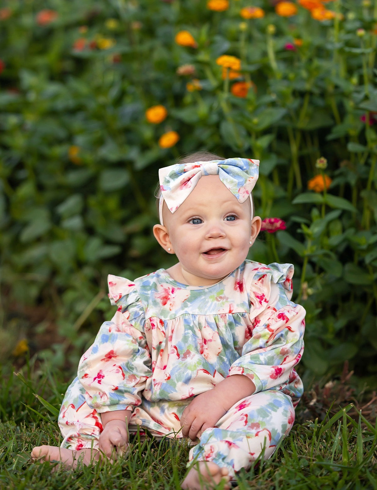 A smiling baby in the Mabel and Honey Floral Flutter Long Sleeve Ruffle Shoulder Bubble Romper, featuring a soft stretch knit fabric with charming floral patterns and a matching bow, sits on green grass as vibrant orange and yellow flowers bloom in the background. The adorable bubble leg design adds extra charm to this delightful scene.