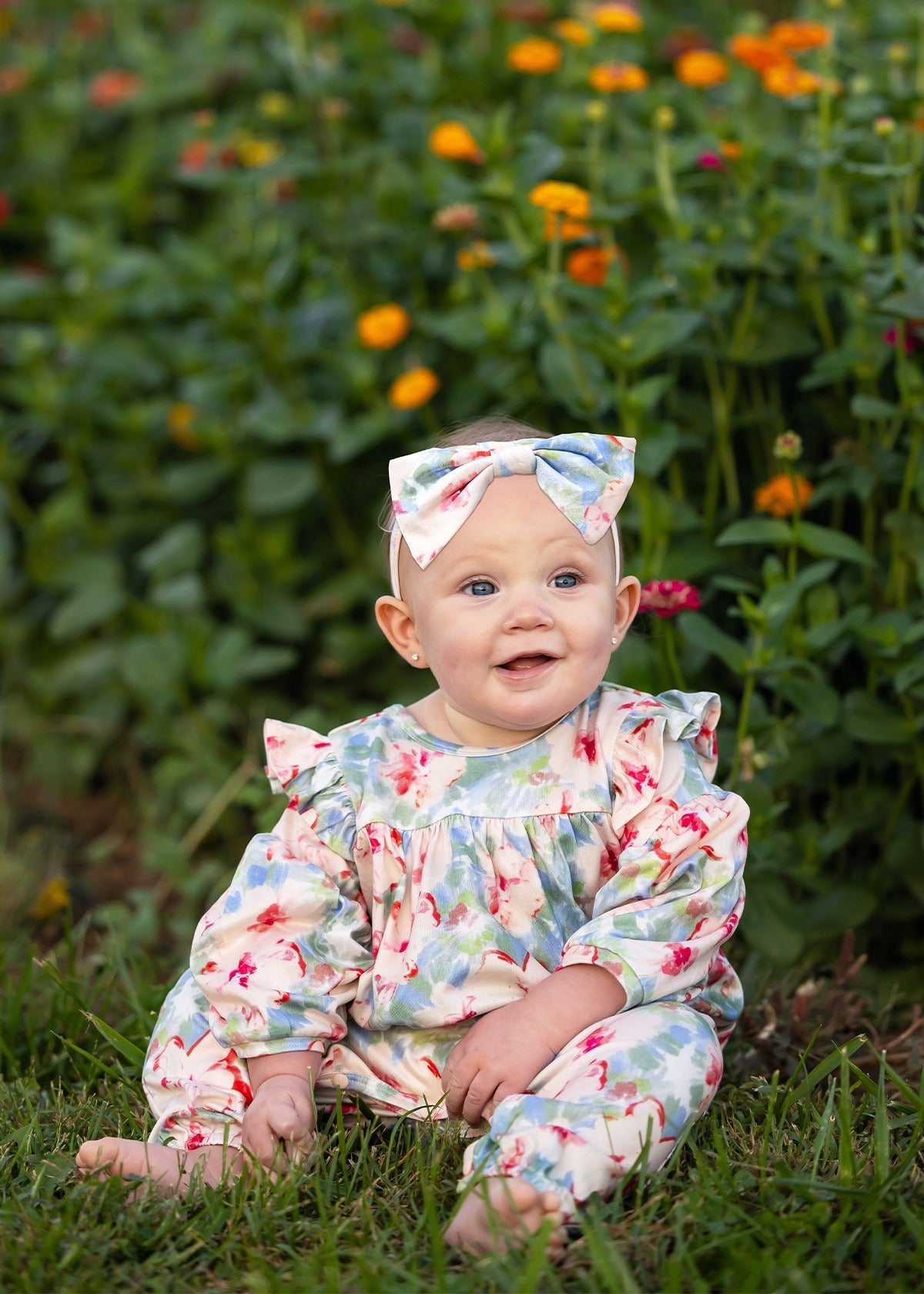 A smiling baby in the Mabel and Honey Floral Flutter Long Sleeve Ruffle Shoulder Bubble Romper, featuring a soft stretch knit fabric with charming floral patterns and a matching bow, sits on green grass as vibrant orange and yellow flowers bloom in the background. The adorable bubble leg design adds extra charm to this delightful scene.