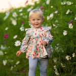 A smiling toddler with blonde hair walks through tall green grass and white wildflowers, wearing the Mabel and Honey Floral Flutter Long Sleeve Ruffle Shoulder Top and Legging Set.
