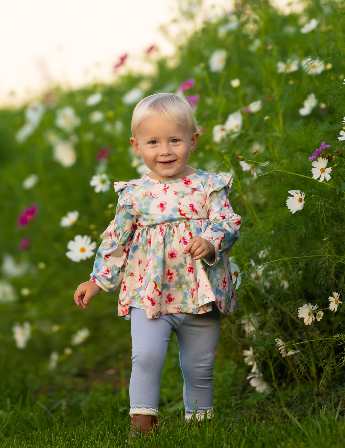 A smiling toddler with blonde hair walks through tall green grass and white wildflowers, wearing the Mabel and Honey Floral Flutter Long Sleeve Ruffle Shoulder Top and Legging Set.