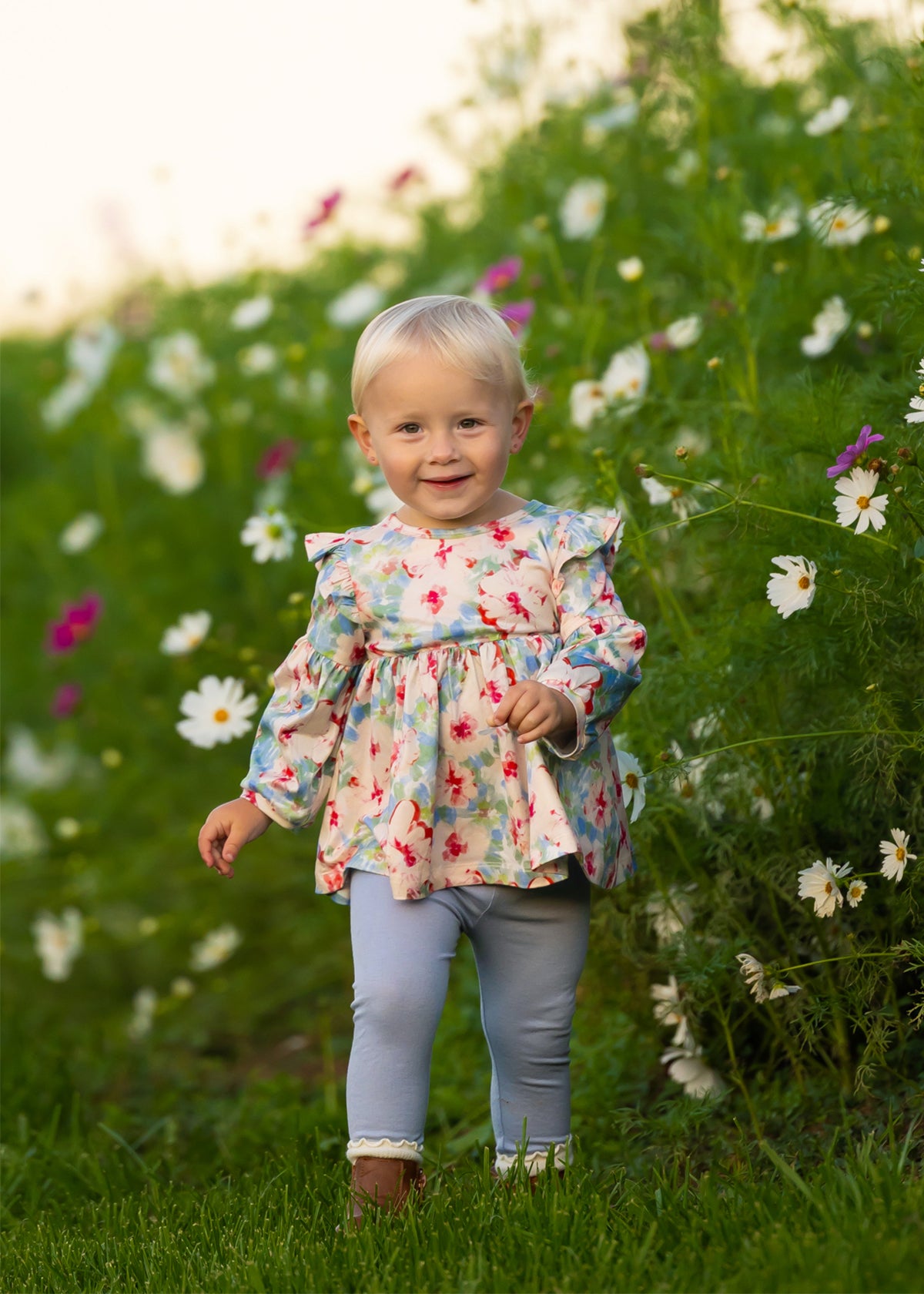 A smiling toddler with blonde hair walks through tall green grass and white wildflowers, wearing the Mabel and Honey Floral Flutter Long Sleeve Ruffle Shoulder Top and Legging Set.