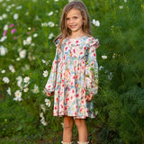 A young girl stands smiling in a garden with tall green plants and white flowers in the background, wearing the Mabel and Honey Floral Flutter Dress, which features long sleeves, ruffle shoulders, and a tiered skirt.
