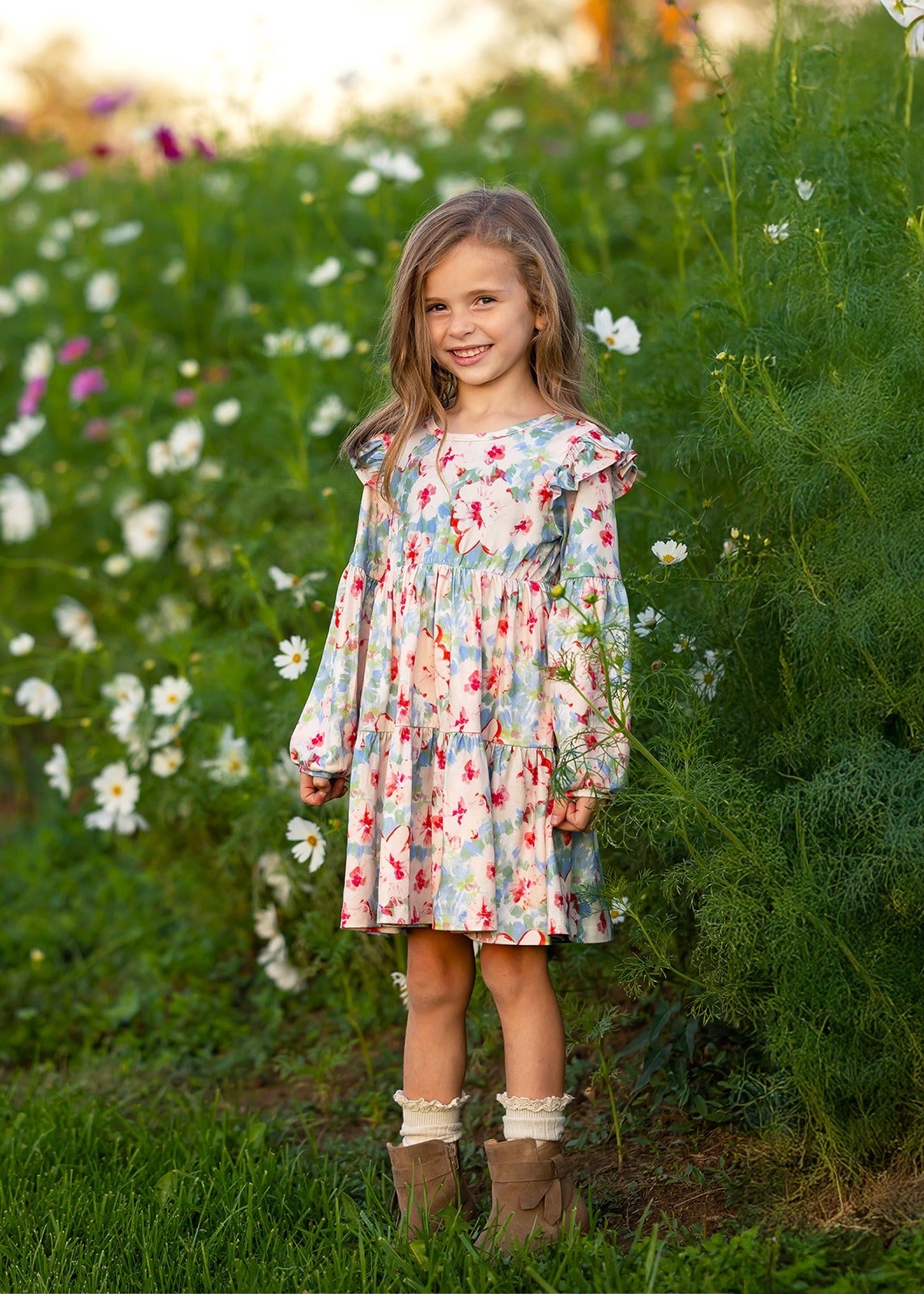 A young girl stands smiling in a garden with tall green plants and white flowers in the background, wearing the Mabel and Honey Floral Flutter Dress, which features long sleeves, ruffle shoulders, and a tiered skirt.