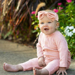 A smiling baby wearing the Mabel and Honey Margret Long Sleeve Solid Flower Button Straight Leg Romper in heathered pink, complete with flower-shaped buttons and a matching headband, sits on a sidewalk surrounded by colorful flowers and lush green plants. The soft knit fabric of the romper adds to the cozy, cheerful scene.