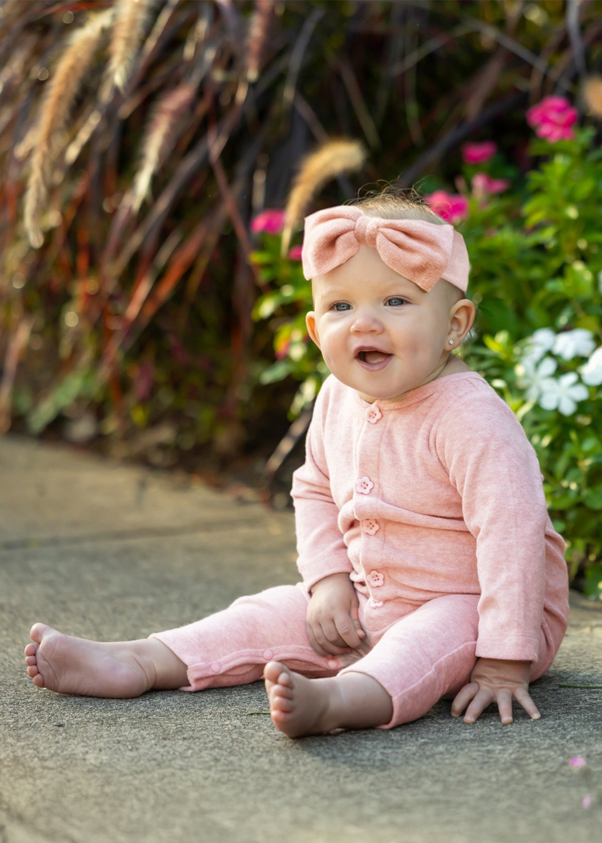 A smiling baby wearing the Mabel and Honey Margret Long Sleeve Solid Flower Button Straight Leg Romper in heathered pink, complete with flower-shaped buttons and a matching headband, sits on a sidewalk surrounded by colorful flowers and lush green plants. The soft knit fabric of the romper adds to the cozy, cheerful scene.