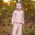 A young child stands and smiles outdoors among green and brown foliage, wearing the Mabel and Honey Margret Long Sleeve Solid Peplum Ruffle Hem Top and Pant Set in heathered pink. The charming ensemble features a peplum ruffle hem top paired with matching pants, adding a sweet touch to the look. Grass and trees provide a natural background.