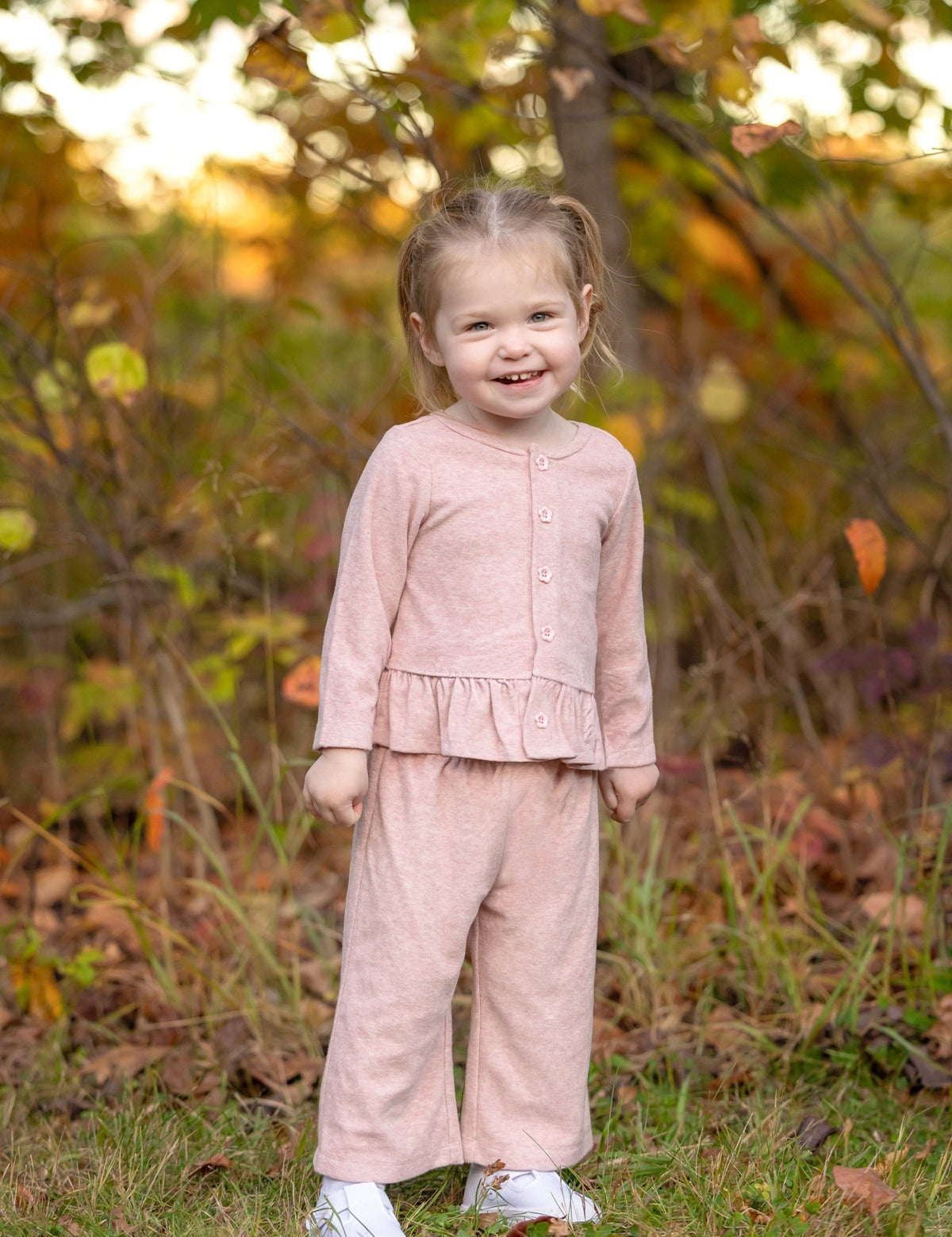 A young child stands and smiles outdoors among green and brown foliage, wearing the Mabel and Honey Margret Long Sleeve Solid Peplum Ruffle Hem Top and Pant Set in heathered pink. The charming ensemble features a peplum ruffle hem top paired with matching pants, adding a sweet touch to the look. Grass and trees provide a natural background.
