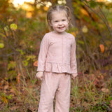 A young child stands and smiles outdoors among green and brown foliage, wearing the Mabel and Honey Margret Long Sleeve Solid Peplum Ruffle Hem Top and Pant Set in heathered pink. The charming ensemble features a peplum ruffle hem top paired with matching pants, adding a sweet touch to the look. Grass and trees provide a natural background.